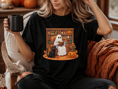 May include: A black t-shirt featuring a graphic print of a white ghost sitting in an orange armchair, reading a book and holding a mug. A black cat sits nearby on a rug with autumn leaves, next to a jack-o'-lantern and a wooden bookshelf filled with books.
