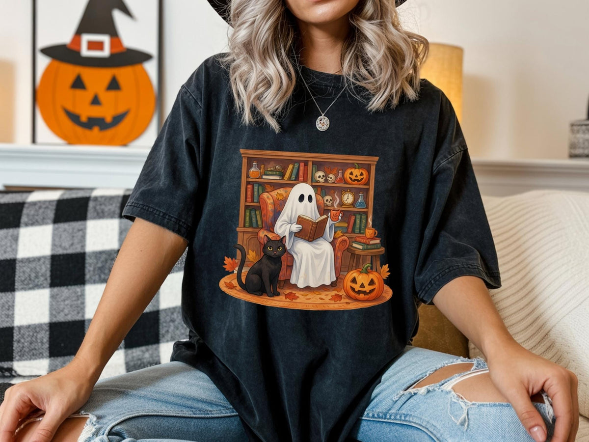 May include: A black short-sleeve t-shirt featuring a graphic print of a white ghost sitting in an orange armchair reading a book. A black cat sits on the floor nearby. The scene includes a wooden bookshelf, pumpkins, and autumn leaves.