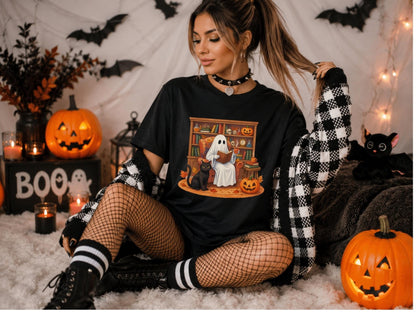 May include: A black t-shirt featuring a graphic print of a white ghost sitting in an armchair reading a book, accompanied by a black cat and pumpkins in front of a bookshelf. The design includes autumn leaves on the floor and various small items.