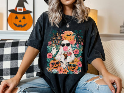 May include: A black short-sleeve t-shirt featuring a colorful graphic of a ghost wearing sunglasses and blowing a pink bubble, holding a coconut drink. The design includes two jack-o'-lanterns, tropical flowers, palm trees, and a sunset.