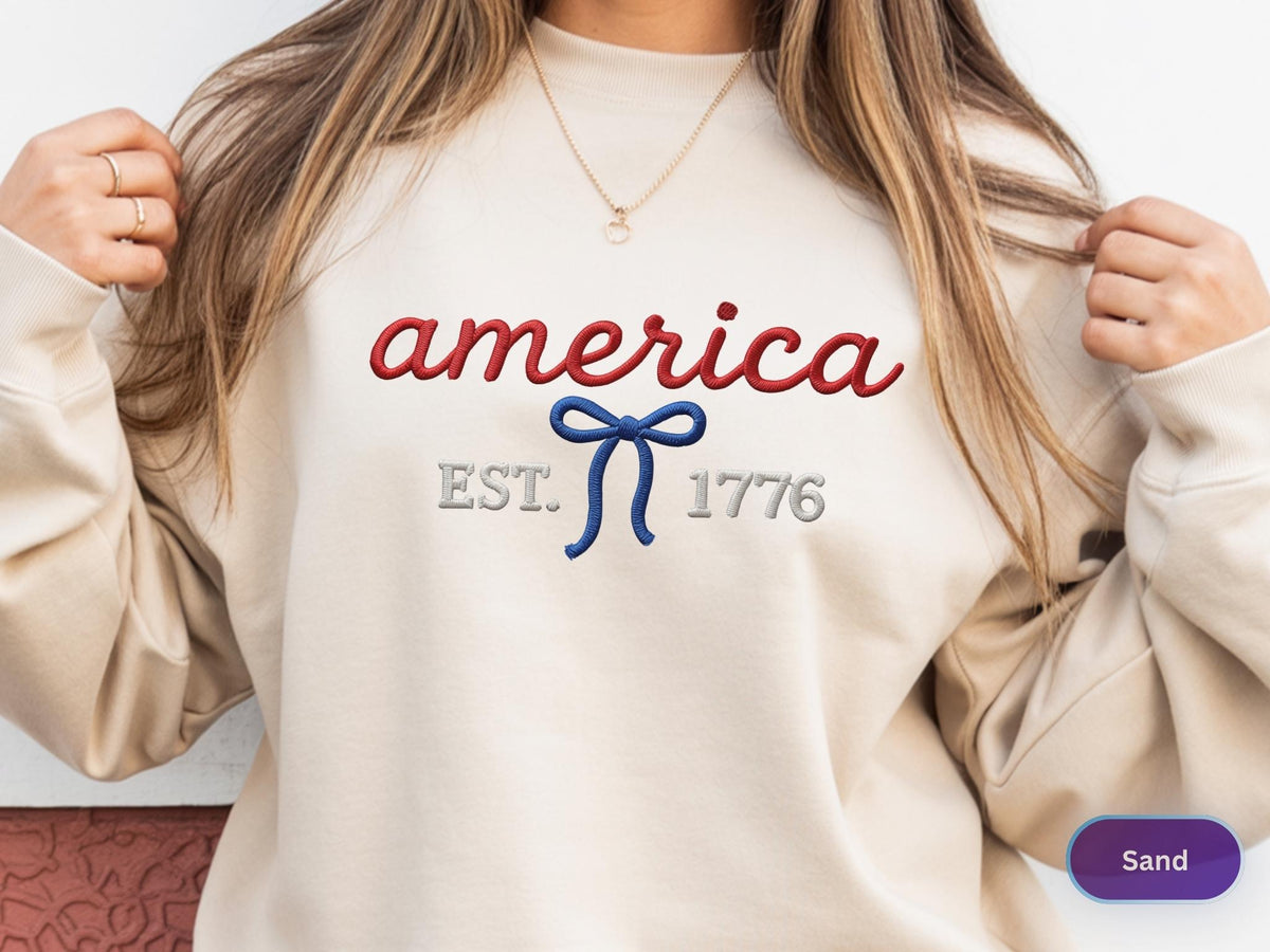 May include: A sand-colored crewneck sweatshirt featuring red embroidered text that reads america, a blue embroidered bow graphic, and grey embroidered text that reads EST. 1776. The design is centered on the chest of the long-sleeved garment.