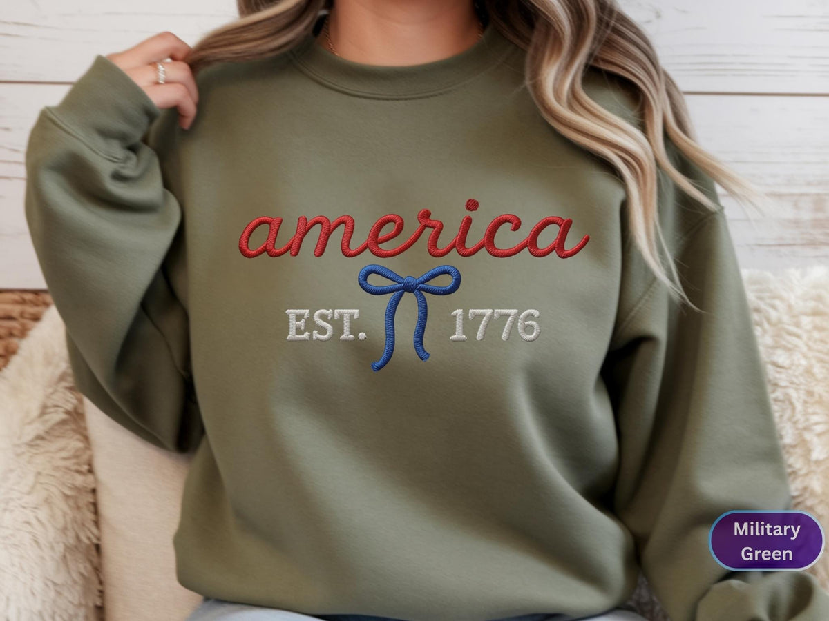 May include: A military green crewneck sweatshirt featuring red embroidered cursive text that reads america, a blue embroidered bow graphic, and white embroidered text that reads EST. 1776 centered on the chest.