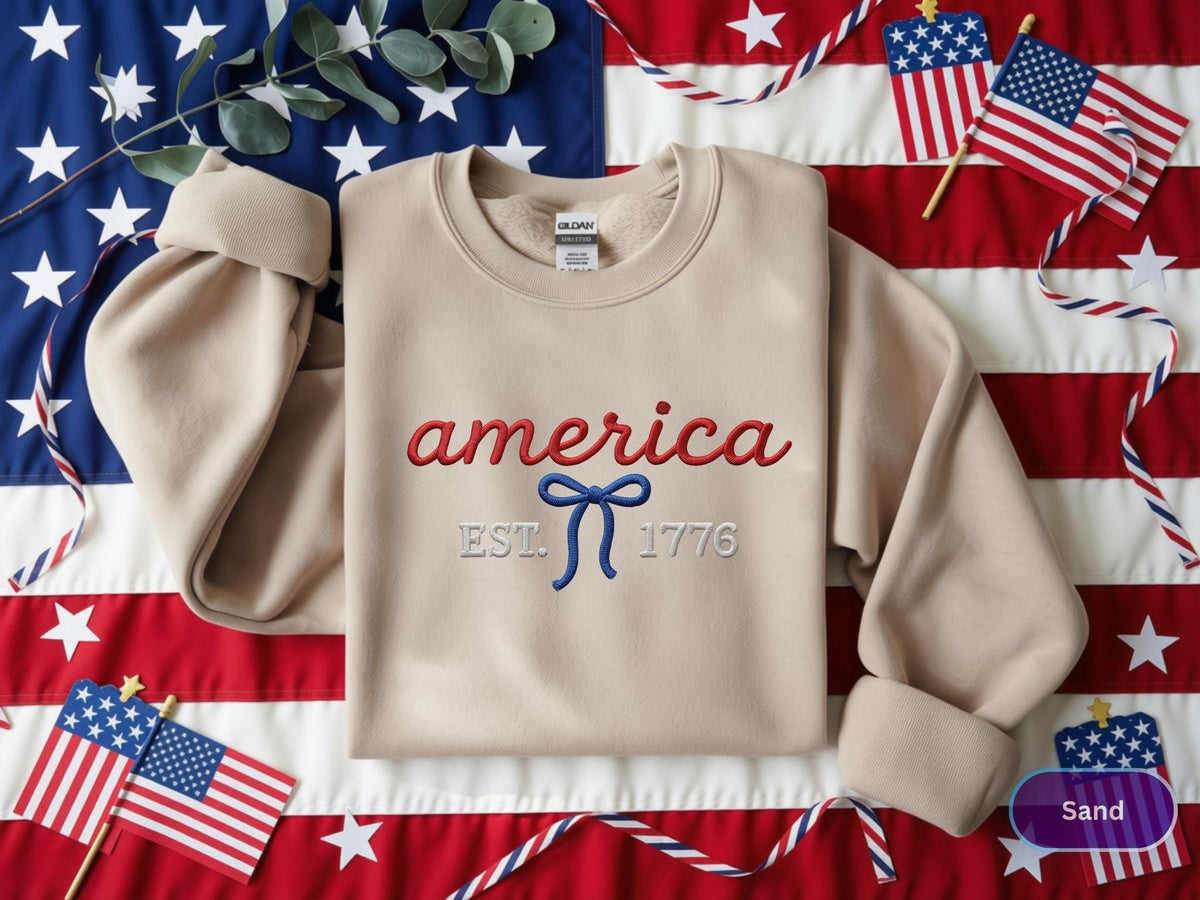 May include: A sand-colored crewneck sweatshirt featuring red embroidered text that reads america, a blue embroidered bow graphic, and white embroidered text that reads EST. 1776 centered on the chest. The garment has long sleeves and a ribbed collar.