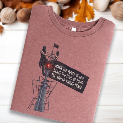 May include: A mauve crew neck t-shirt featuring a graphic of two figures on a tower holding a black flag with the text: When the power of love beats the love of power the world knows peace. The design includes a small red light on the tower structure.