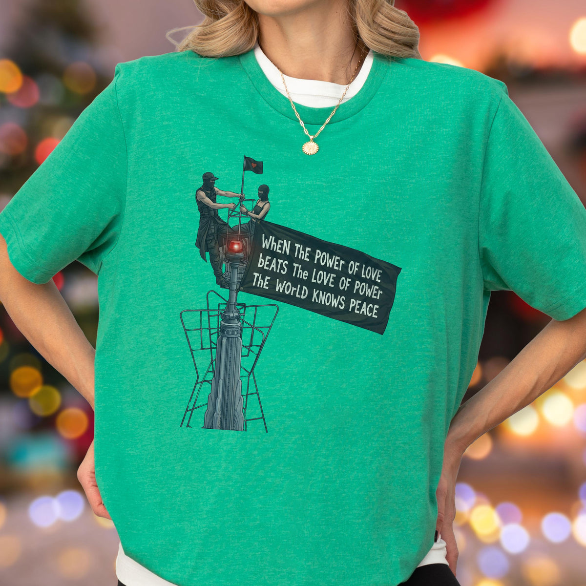 May include: A bright green short-sleeved t-shirt featuring a graphic illustration of two figures atop a structure holding a banner that reads, "WHEN THE POWER OF LOVE BEATS THE LOVE OF POWER THE WORLD KNOWS PEACE." A gold chain necklace is worn.