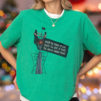 May include: A bright green short-sleeved t-shirt featuring a graphic illustration of two figures atop a structure holding a banner that reads, "WHEN THE POWER OF LOVE BEATS THE LOVE OF POWER THE WORLD KNOWS PEACE." A gold chain necklace is worn.