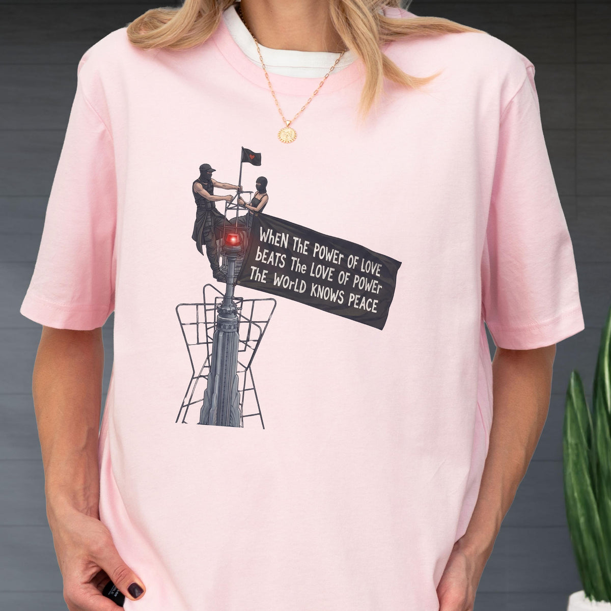 May include: A light pink short-sleeved t-shirt featuring a graphic of two figures atop a structure holding a black banner. The banner displays the text: WHEN THE POWER OF LOVE BEATS THE LOVE OF POWER THE WORLD KNOWS PEACE. A gold chain necklace is visible.