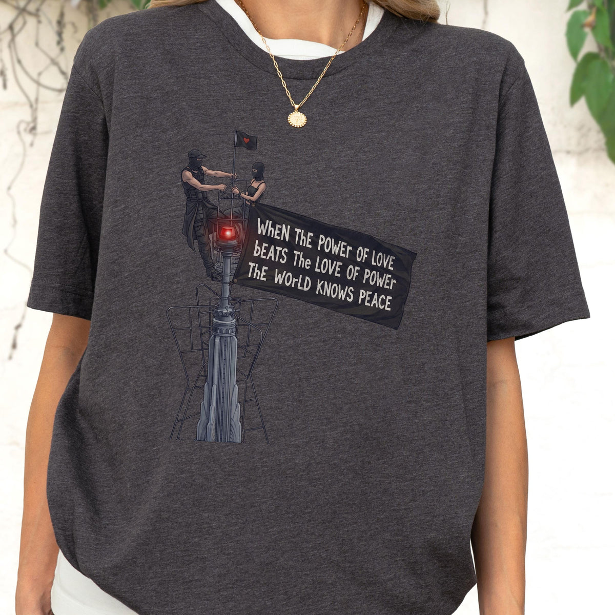 May include: A charcoal gray t-shirt featuring a graphic print of two figures standing atop a tall structure, holding a black flag with the text: WHEN THE POWER OF LOVE BEATS THE LOVE OF POWER THE WORLD KNOWS PEACE. The design includes a glowing red light.