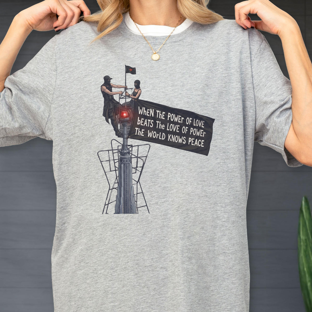 May include: A heather gray t-shirt featuring a graphic of two figures standing atop a tall structure, holding a black flag with the text: WHEN THE POWER OF LOVE BEATS THE LOVE OF POWER THE WORLD KNOWS PEACE. The design includes a small red light.