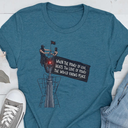 May include: A teal blue short-sleeved t-shirt featuring a graphic of two figures standing atop a tall structure, holding a black flag with the text: WHEN THE POWER OF LOVE BEATS THE LOVE OF POWER THE WORLD KNOWS PEACE. The design includes a red light.