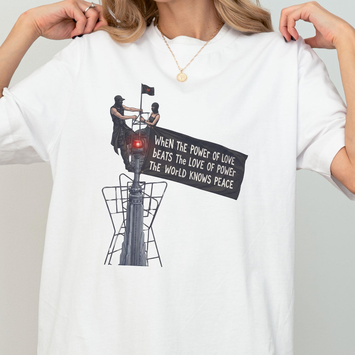 May include: A white t-shirt featuring a graphic illustration of two figures standing atop a tall structure, holding a black flag with the text: WHEN THE POWER OF LOVE BEATS THE LOVE OF POWER THE WORLD KNOWS PEACE. The design includes a red light.