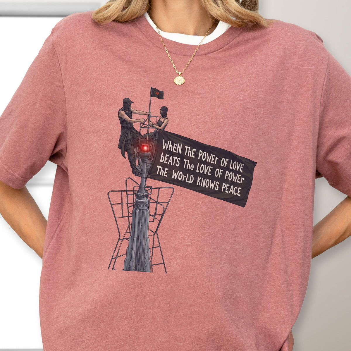 May include: A dusty rose t-shirt featuring a graphic of two figures atop a tall structure holding a black flag. The flag displays the text: WHEN THE POWER OF LOVE BEATS THE LOVE OF POWER THE WORLD KNOWS PEACE. The design includes a small red light.
