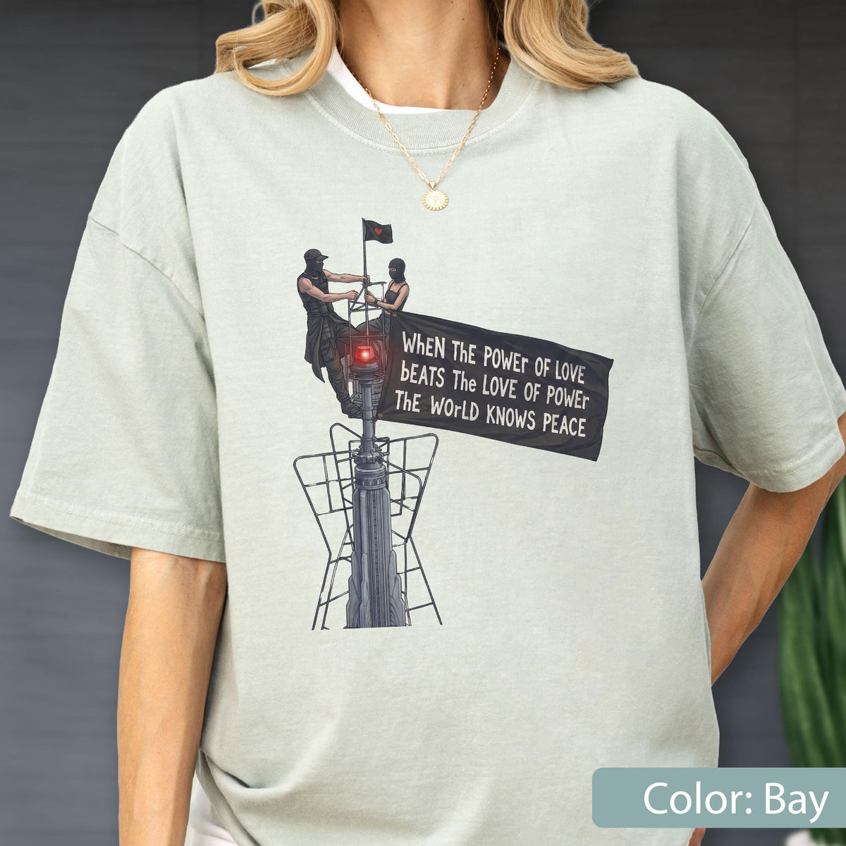 May include: A light sage green t-shirt featuring a graphic print of two figures atop a tall structure holding a black banner. The text on the banner reads: When the power of love beats the love of power the world knows peace. The design includes a red light.
