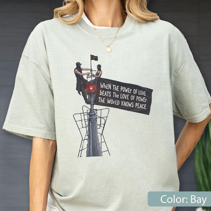 May include: A light sage green t-shirt featuring a graphic print of two figures atop a tall structure holding a black banner. The text on the banner reads: When the power of love beats the love of power the world knows peace. The design includes a red light.