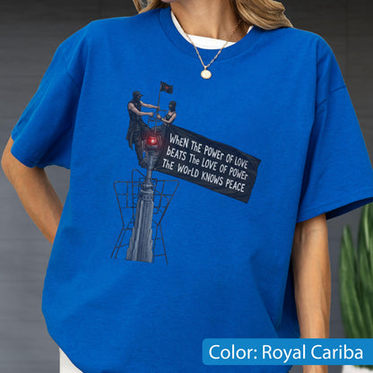 May include: A royal blue short-sleeve t-shirt featuring a graphic print of two figures atop a tower holding a black flag. The flag displays the text: WHEN THE POWER OF LOVE BEATS THE LOVE OF POWER THE WORLD KNOWS PEACE. A red light glows on the tower.