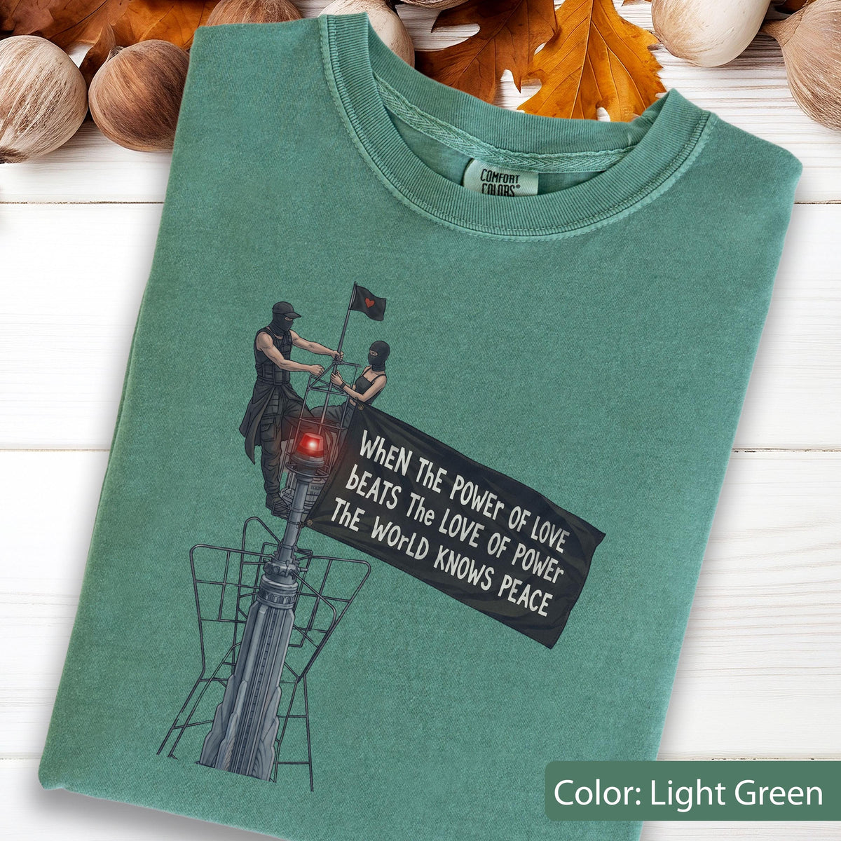 May include: A light green t-shirt featuring a graphic of two figures standing on a tall structure, one holding a black flag with the text: WHEN THE POWER OF LOVE BEATS THE LOVE OF POWER THE WORLD KNOWS PEACE. The shirt has a crew neckline.