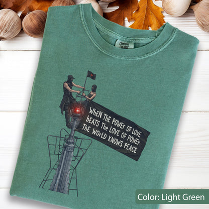 May include: A light green t-shirt featuring a graphic of two figures standing on a tall structure, one holding a black flag with the text: WHEN THE POWER OF LOVE BEATS THE LOVE OF POWER THE WORLD KNOWS PEACE. The shirt has a crew neckline.