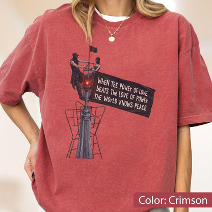 May include: A crimson short-sleeve t-shirt featuring a graphic of two figures atop a tall structure holding a black flag. The flag displays the text: WHEN THE POWER OF LOVE BEATS THE LOVE OF POWER THE WORLD KNOWS PEACE. The shirt has a crew neckline.