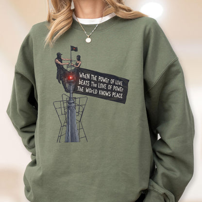 May include: An olive green crewneck sweatshirt featuring a graphic print of two figures atop a tall structure. They hold a black flag with the text: When the power of love beats the love of power the world knows peace. The design includes a red light.