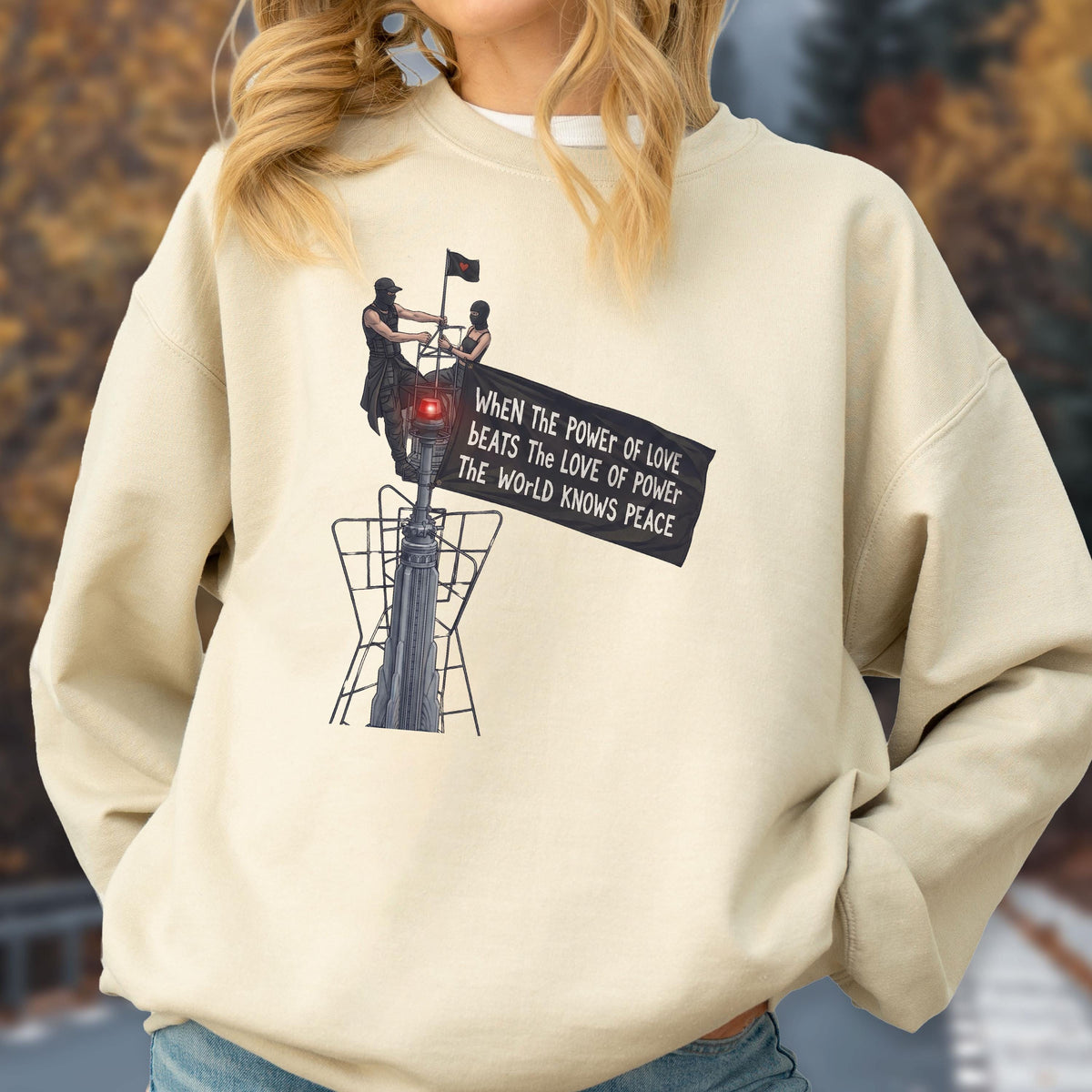 May include: A beige crewneck sweatshirt featuring a graphic print of two figures atop a tall structure holding a black flag. The flag displays the text: When the power of love beats the love of power the world knows peace. The design includes a red light.