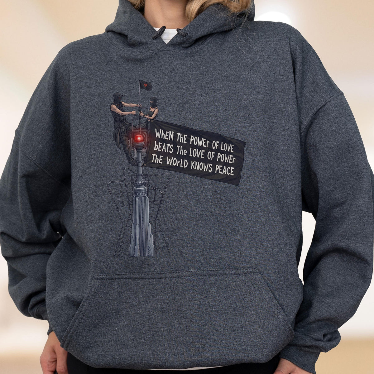 May include: A dark heather gray hooded sweatshirt featuring a graphic print of two figures atop a tall structure. The design includes a black flag with the text: When the power of love beats the love of power the world knows peace.