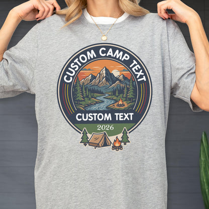 May include: A heather gray t-shirt featuring a circular graphic with a mountain landscape, a river, and a campfire. The text reads CUSTOM CAMP TEXT at the top and bottom, with the year 2026 below. A small tent and trees are depicted at the base.