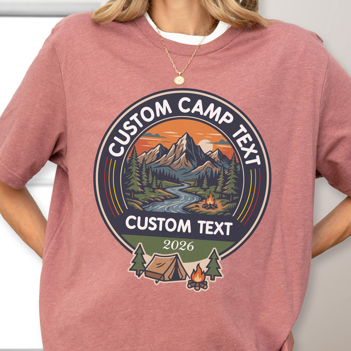 May include: A dusty rose short-sleeve t-shirt featuring a circular graphic with a mountain landscape, a river, a campfire, and a tent. The text reads CUSTOM CAMP TEXT at the top and bottom, with the year 2026 printed in the center of the lower section.