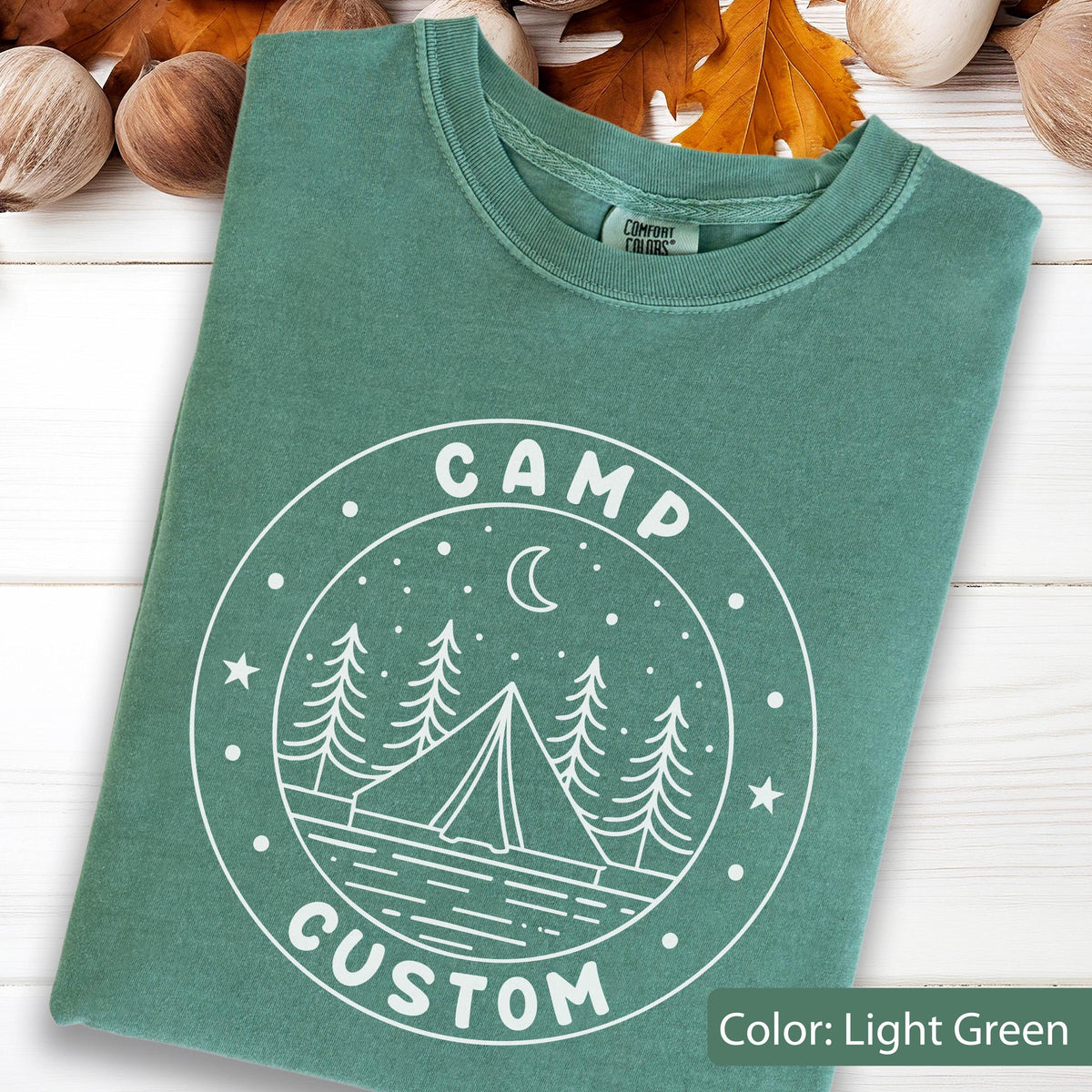 May include: A light green t-shirt featuring a white circular graphic print. The design includes a tent, pine trees, a crescent moon, and stars, with the text "CAMP" at the top and "CUSTOM" at the bottom. The shirt has a crew neckline and a Comfort Colors label.