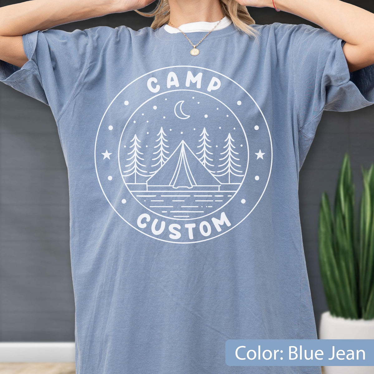 May include: A blue jean colored short-sleeve t-shirt featuring a white circular graphic print. The design includes a tent, pine trees, a crescent moon, stars, and the text CAMP CUSTOM arranged within the circular border of the graphic.