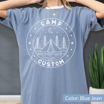 May include: A blue jean colored short-sleeve t-shirt featuring a white circular graphic print. The design includes a tent, pine trees, a crescent moon, stars, and the text CAMP CUSTOM arranged within the circular border of the graphic.