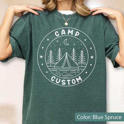 May include: A blue spruce short-sleeve t-shirt featuring a white circular graphic on the chest. The design depicts a tent, pine trees, a crescent moon, and stars, with the text CAMP CUSTOM printed in a circular border around the illustration.