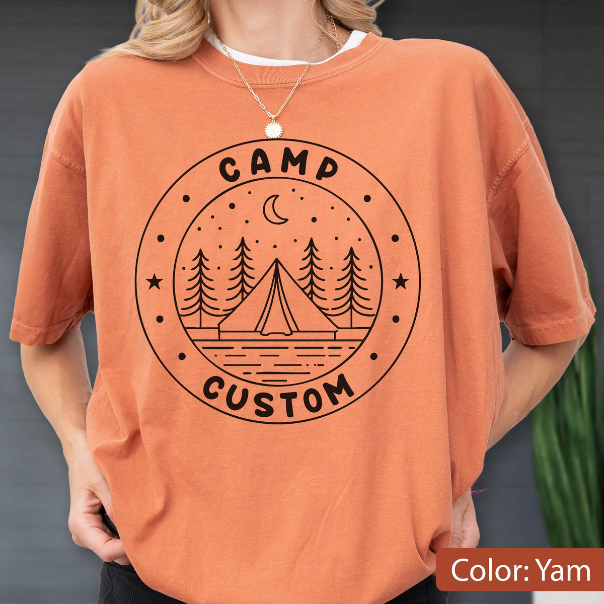 May include: A yam-colored short-sleeve t-shirt featuring a black circular graphic on the chest. The design includes a tent, pine trees, a crescent moon, and stars, with the text CAMP CUSTOM printed in a bold, sans-serif font within the circle.