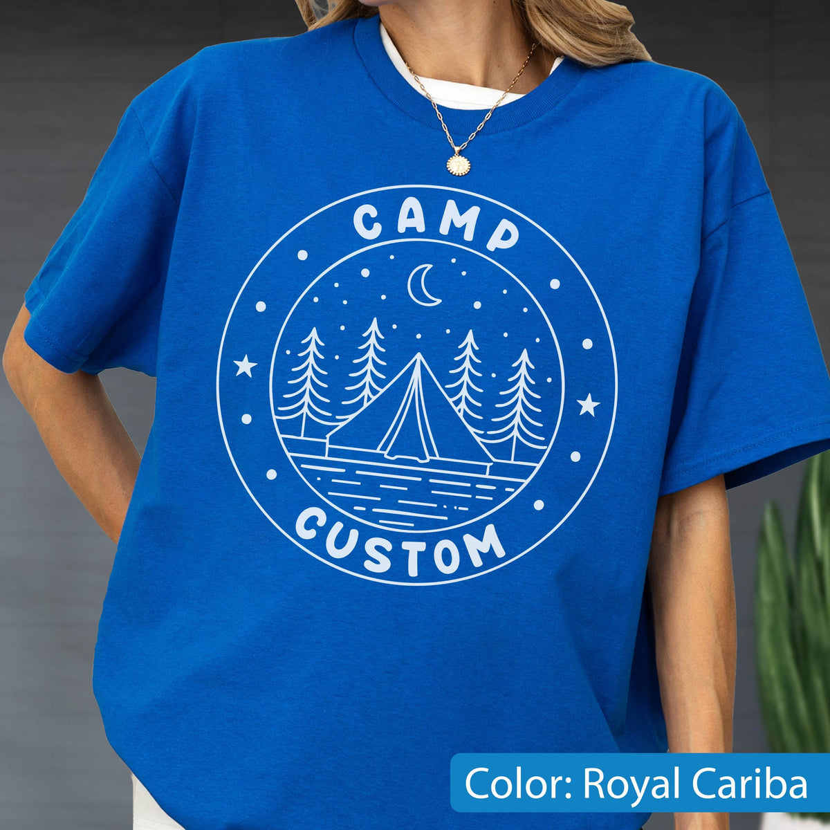 May include: A royal blue t-shirt featuring a white circular graphic print. The design includes a tent, pine trees, a crescent moon, and stars, with the text CAMP CUSTOM arched around the border. The shirt has short sleeves and a crew neckline.