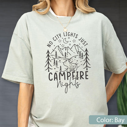 May include: A light sage green short-sleeve t-shirt featuring a black graphic print of a mountain landscape, pine trees, a crescent moon, stars, and a campfire. The text reads NO CITY LIGHTS JUST CAMPFIRE Nights in a mix of block and script fonts.