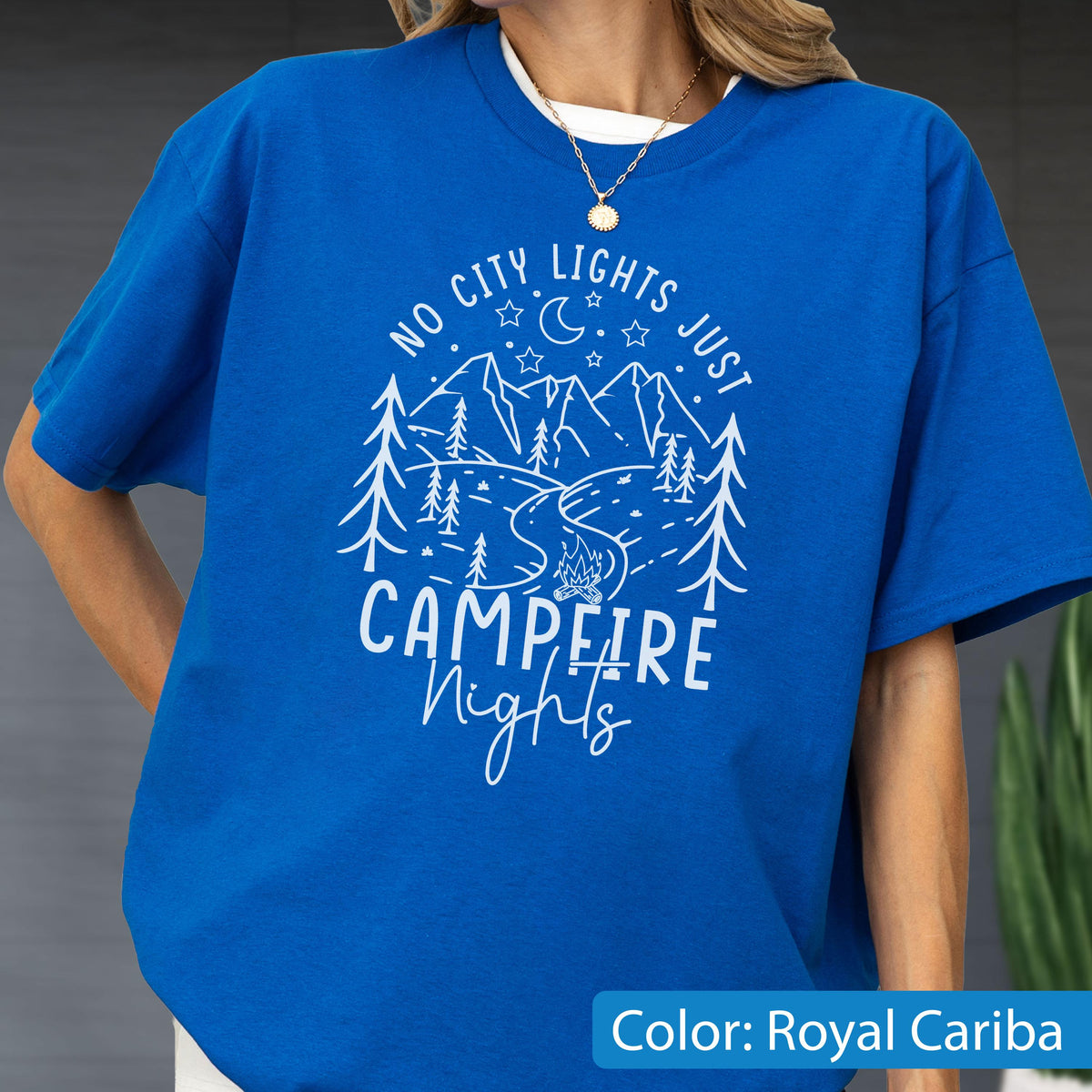 May include: A royal blue short-sleeve t-shirt featuring a white graphic print of mountains, pine trees, a crescent moon, stars, and a campfire. The text reads NO CITY LIGHTS JUST CAMPFIRE Nights in a mix of block and script lettering.