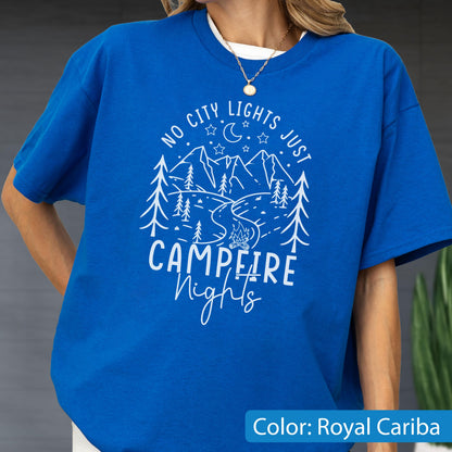 May include: A royal blue short-sleeve t-shirt featuring a white graphic print of mountains, pine trees, a crescent moon, stars, and a campfire. The text reads NO CITY LIGHTS JUST CAMPFIRE Nights in a mix of block and script lettering.
