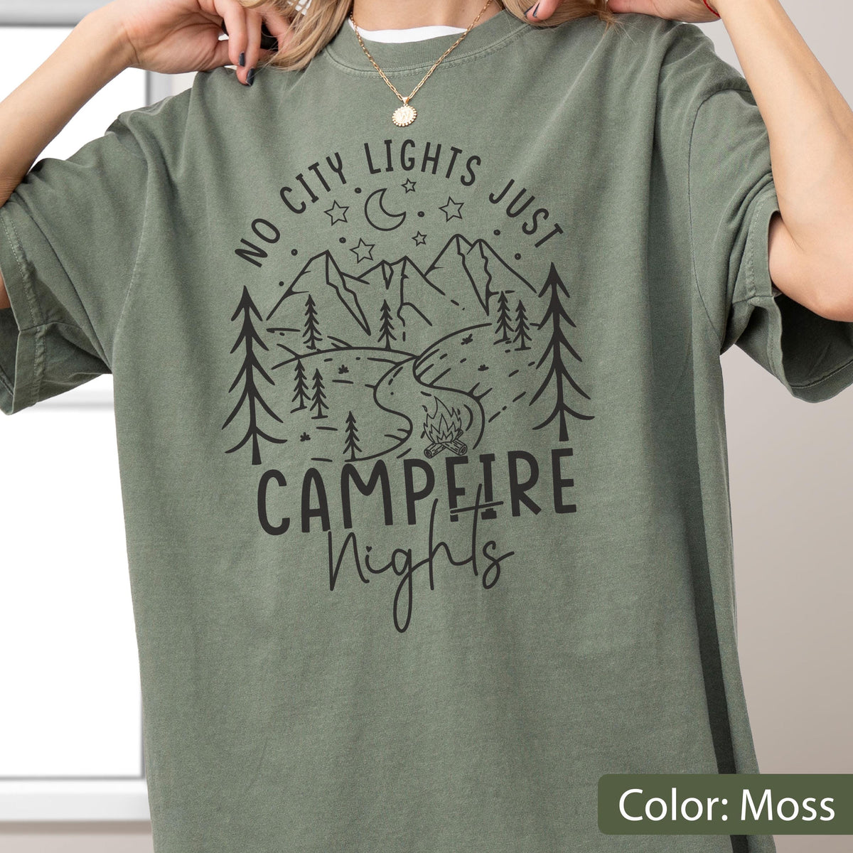 May include: A moss green short-sleeve t-shirt featuring a black graphic print of mountains, pine trees, a winding path, a campfire, and stars with a crescent moon. The text reads NO CITY LIGHTS JUST CAMPFIRE NIGHTS in a mix of fonts.