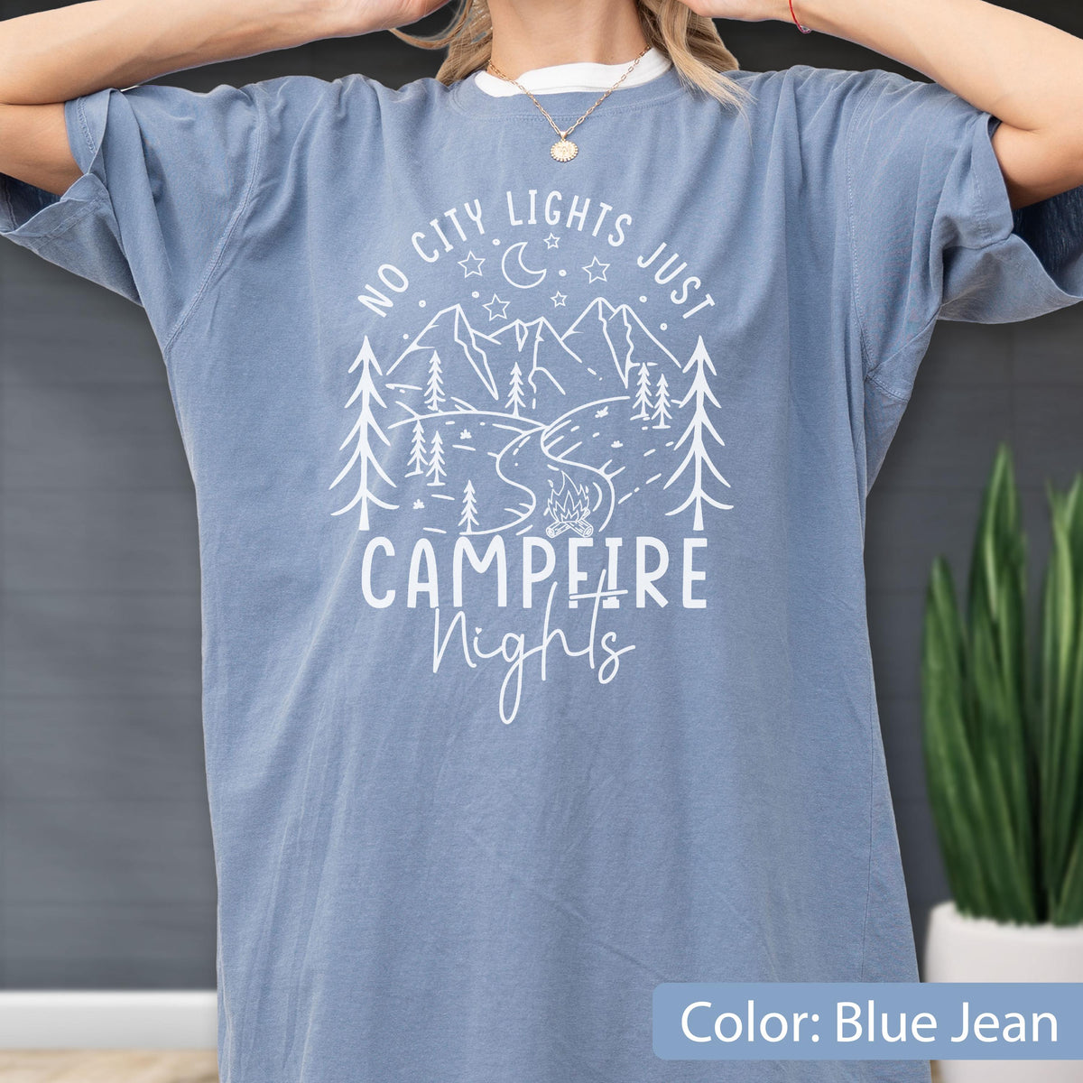 May include: A blue jean colored short-sleeve t-shirt featuring a white graphic print of mountains, pine trees, a crescent moon, stars, and a campfire. The text reads NO CITY LIGHTS JUST CAMPFIRE NIGHTS in a mix of block and script lettering.