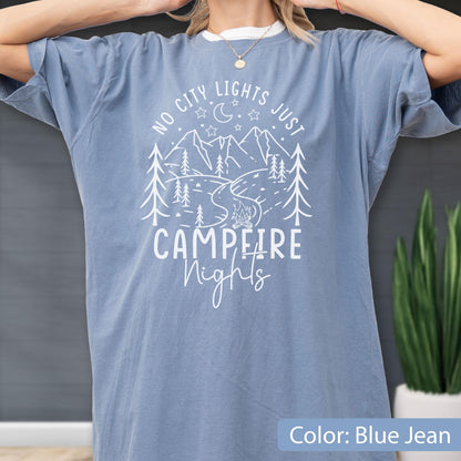 May include: A blue jean colored short-sleeve t-shirt featuring a white graphic print of mountains, pine trees, a crescent moon, stars, and a campfire. The text reads NO CITY LIGHTS JUST CAMPFIRE NIGHTS in a mix of block and script lettering.