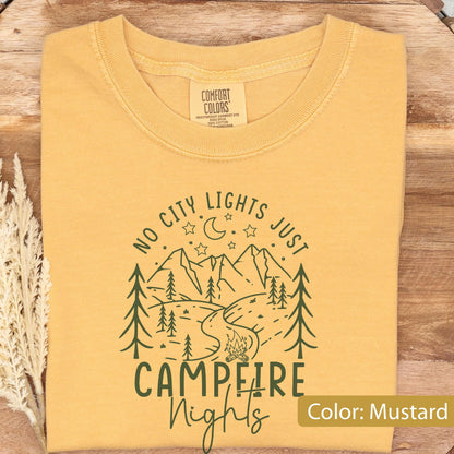May include: A mustard yellow crew neck t-shirt featuring a dark green graphic print of mountains, pine trees, a campfire, and a crescent moon with stars. The text reads NO CITY LIGHTS JUST CAMPFIRE NIGHTS. The shirt has a Comfort Colors brand label.