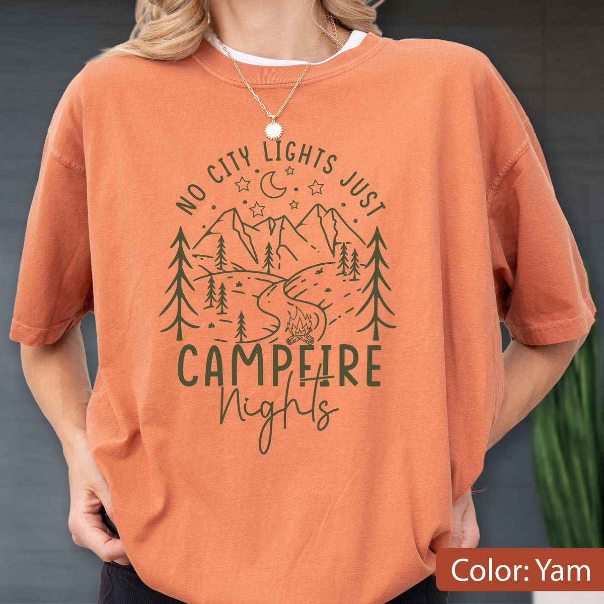 May include: A burnt orange t-shirt featuring a dark green graphic of a mountain landscape, pine trees, a campfire, a crescent moon, and stars. The text reads NO CITY LIGHTS JUST CAMPFIRE Nights in a mix of block and script lettering.