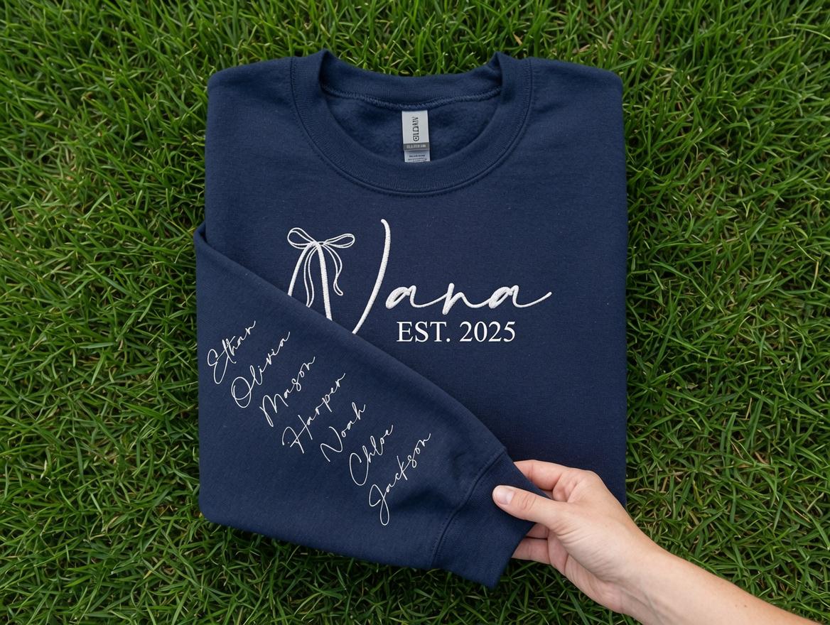May include: A navy blue crewneck sweatshirt featuring white screen-printed text that reads "Nana EST. 2025" with a bow graphic. The sleeve displays a vertical list of names in white cursive script, including Ethan, Olivia, Mason, Harper, Noah, Chloe, and Jackson.