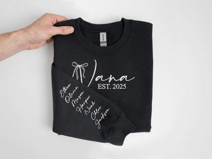 May include: A black long-sleeve sweatshirt featuring white embroidered text that reads "Nana EST. 2025" with a bow graphic. The sleeve displays a vertical list of names including Ethan, Olivia, Mason, Harper, Noah, Chloe, and Jackson in white script.