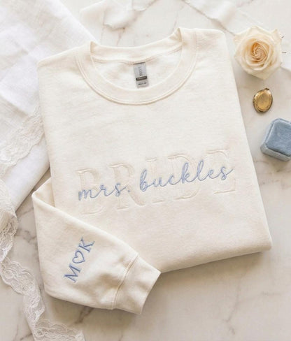 May include: A cream-colored crewneck sweatshirt featuring the word BRIDE embroidered in a large, tonal font across the chest, with the text mrs. buckles in light blue script layered over it. The sleeve features the embroidered letters M, a heart, and K.