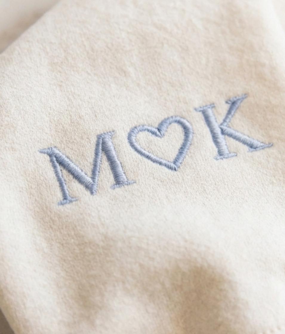 May include: A beige fabric surface featuring light blue embroidery of the letters M and K separated by a heart symbol. The stitching is clearly visible, creating a textured appearance on the soft, neutral-colored textile material.