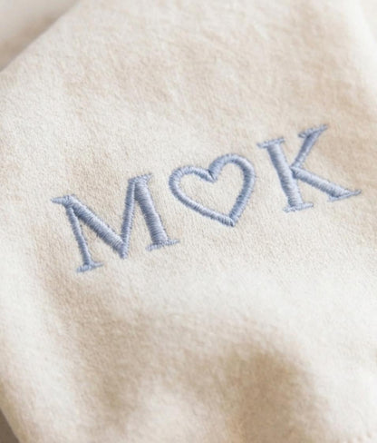 May include: A beige fabric surface featuring light blue embroidery of the letters M and K separated by a heart symbol. The stitching is clearly visible, creating a textured appearance on the soft, neutral-colored textile material.
