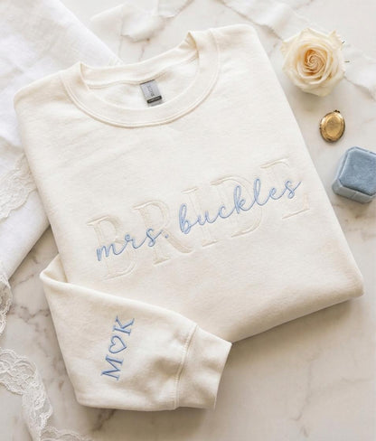 May include: A cream-colored crewneck sweatshirt featuring large, tone-on-tone embroidered block letters spelling BRIDE, with the text mrs. buckles embroidered in light blue script over it. The sleeve features the blue embroidered letters M, a heart, and K.