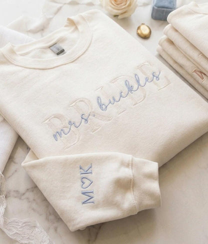 May include: A cream-colored crewneck sweatshirt featuring large, tone-on-tone embroidered block letters and the light blue cursive text "mrs. buckles" across the chest. The sleeve is embroidered with the light blue letters "M" and "K" with a heart.