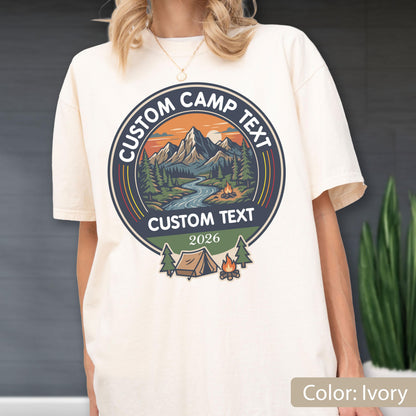 May include: An ivory t-shirt featuring a circular graphic with a mountain landscape, a river, and a campfire. The text reads CUSTOM CAMP TEXT, CUSTOM TEXT, and 2026. Below the circle is a tent and a campfire illustration with two pine trees.