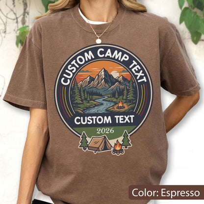 May include: An espresso-colored t-shirt featuring a circular graphic with a mountain landscape, a river, pine trees, a campfire, and a tent. The text reads CUSTOM CAMP TEXT at the top and bottom, with the year 2026 printed below the central design.